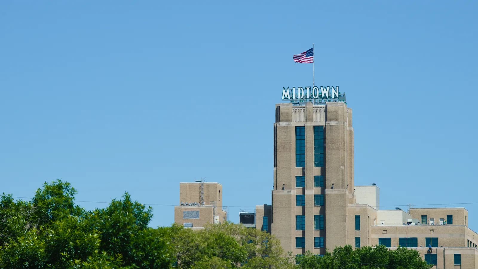 The Midtown Exchange tower with its MIDTOWN sign and a flag on top, rising above the trees in Phillips under a clear blue sky.