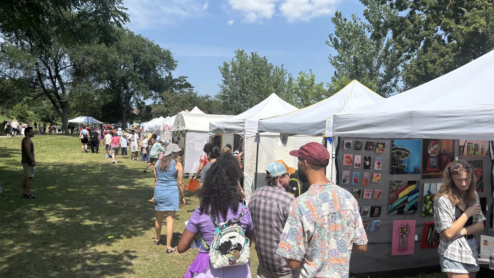 People browsing a long row of white artist tents at the Powderhorn Art Fair in Powderhorn Park on a sunny summer day.