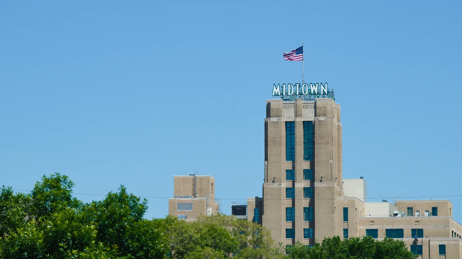 The Midtown Exchange tower with its MIDTOWN sign and a flag on top, rising above the trees in Phillips under a clear blue sky.
