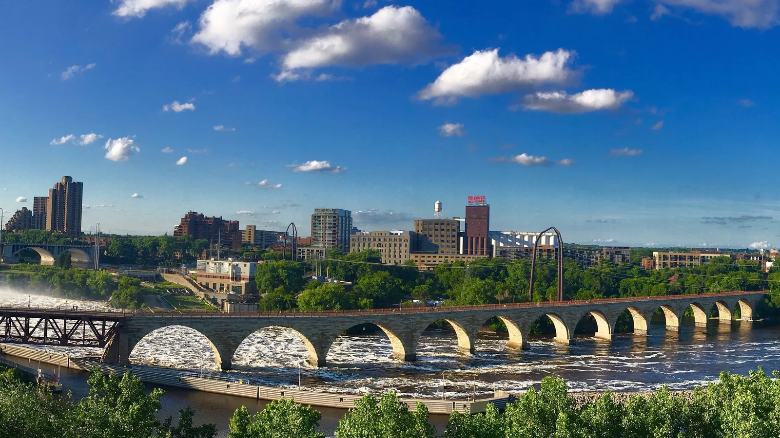 The Stone Arch Bridge over St. Anthony Falls on a sunny summer day, with the St. Anthony Main skyline and the red Pillsbury's Best Flour sign across the Mississippi.