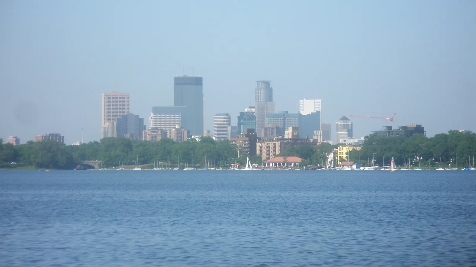 The downtown Minneapolis skyline seen across Bde Maka Ska, with sailboats moored along the far shore and the lake pavilion.