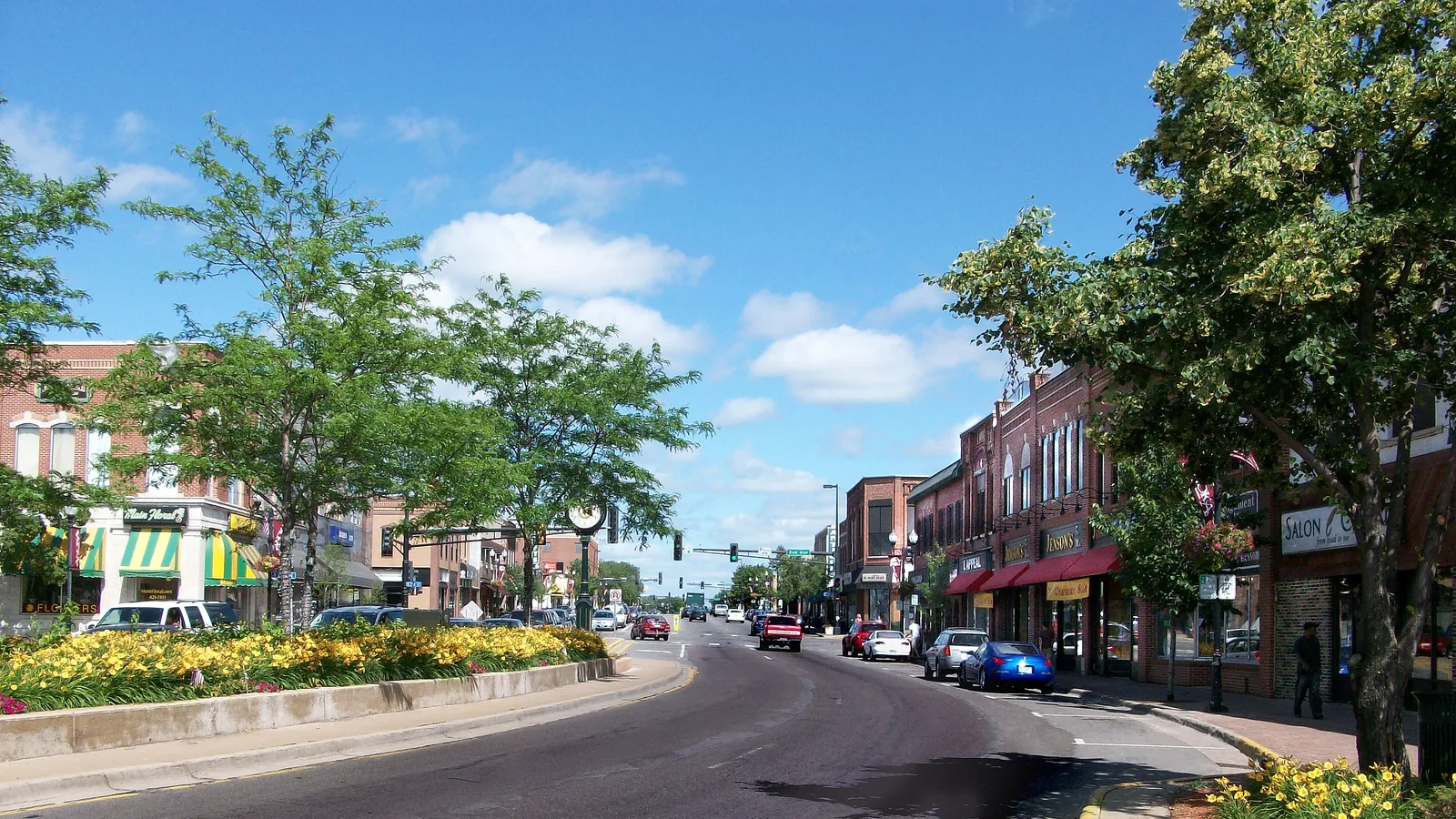 Main Street in downtown Anoka on a sunny summer day, with brick storefronts, red awnings, and a planter full of yellow daylilies.