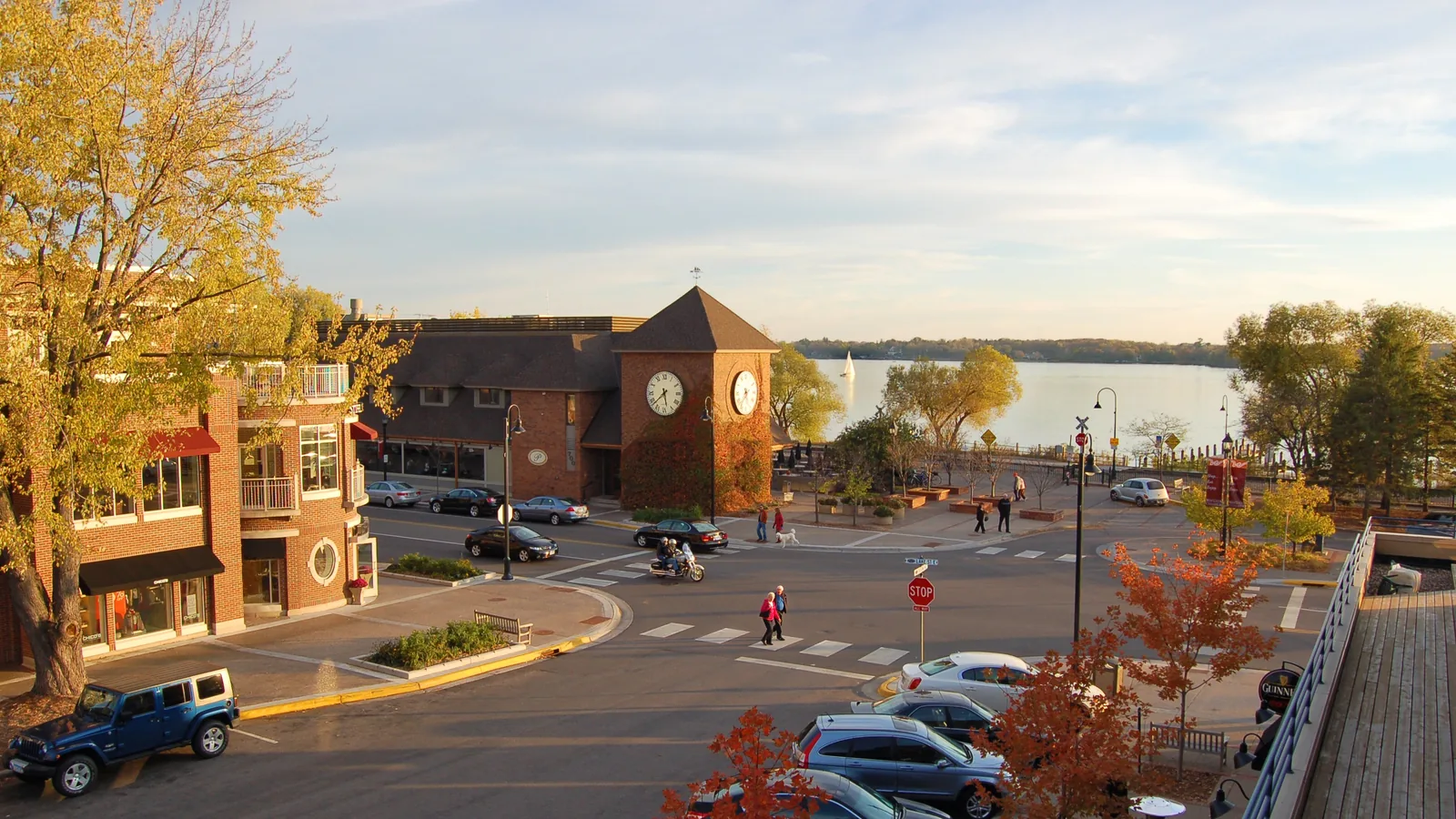 Lake Street in downtown Wayzata on a sunny fall afternoon: the ivy-covered brick clock tower, people crossing the street with a dog, and a sailboat on Wayzata Bay behind it.